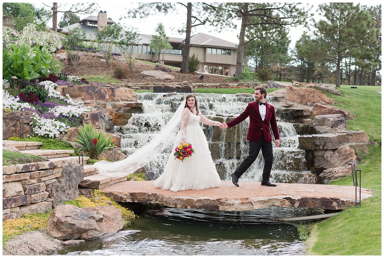 A groom in a red velvet jacket walks with his new bride accross a large rock bridge over a pond at the Cielo at Castle Pines golf course, a castle rock wedding venue