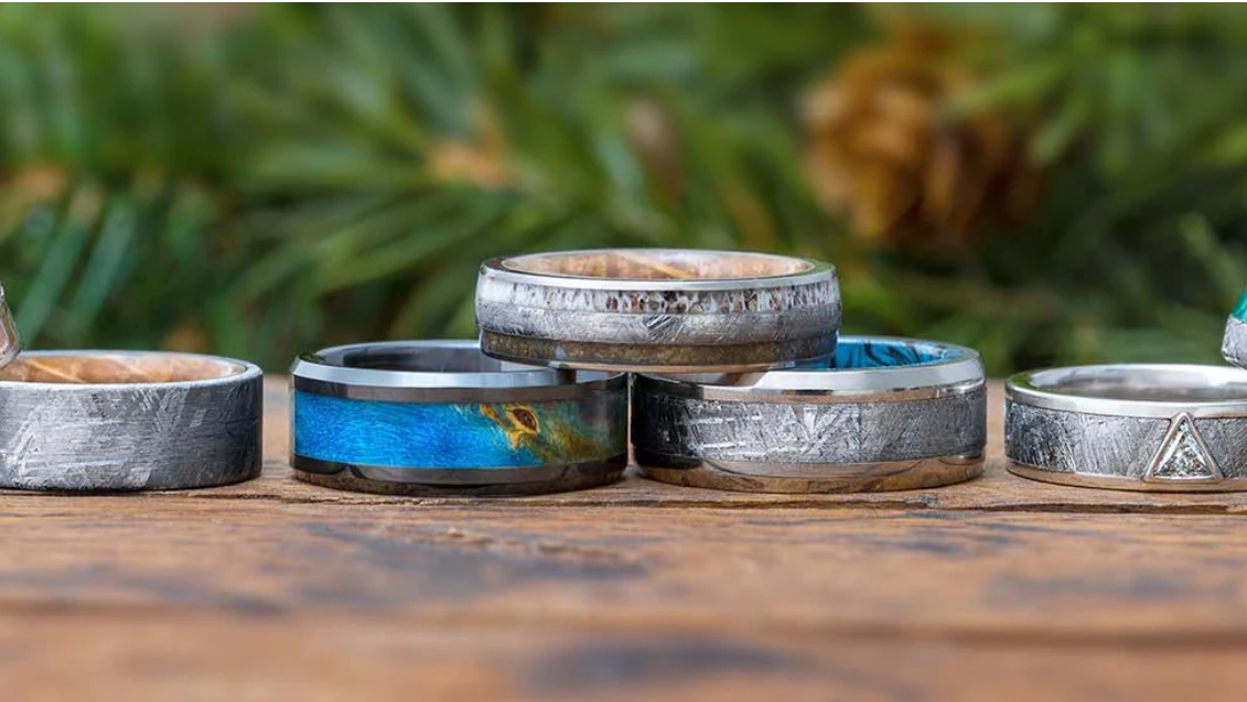 A close up photo of 5 rustic looking men's wedding bands on a wooden table each with different characteristics. One ring is blue with a black outline and the others are all silver with varying rustic features. 