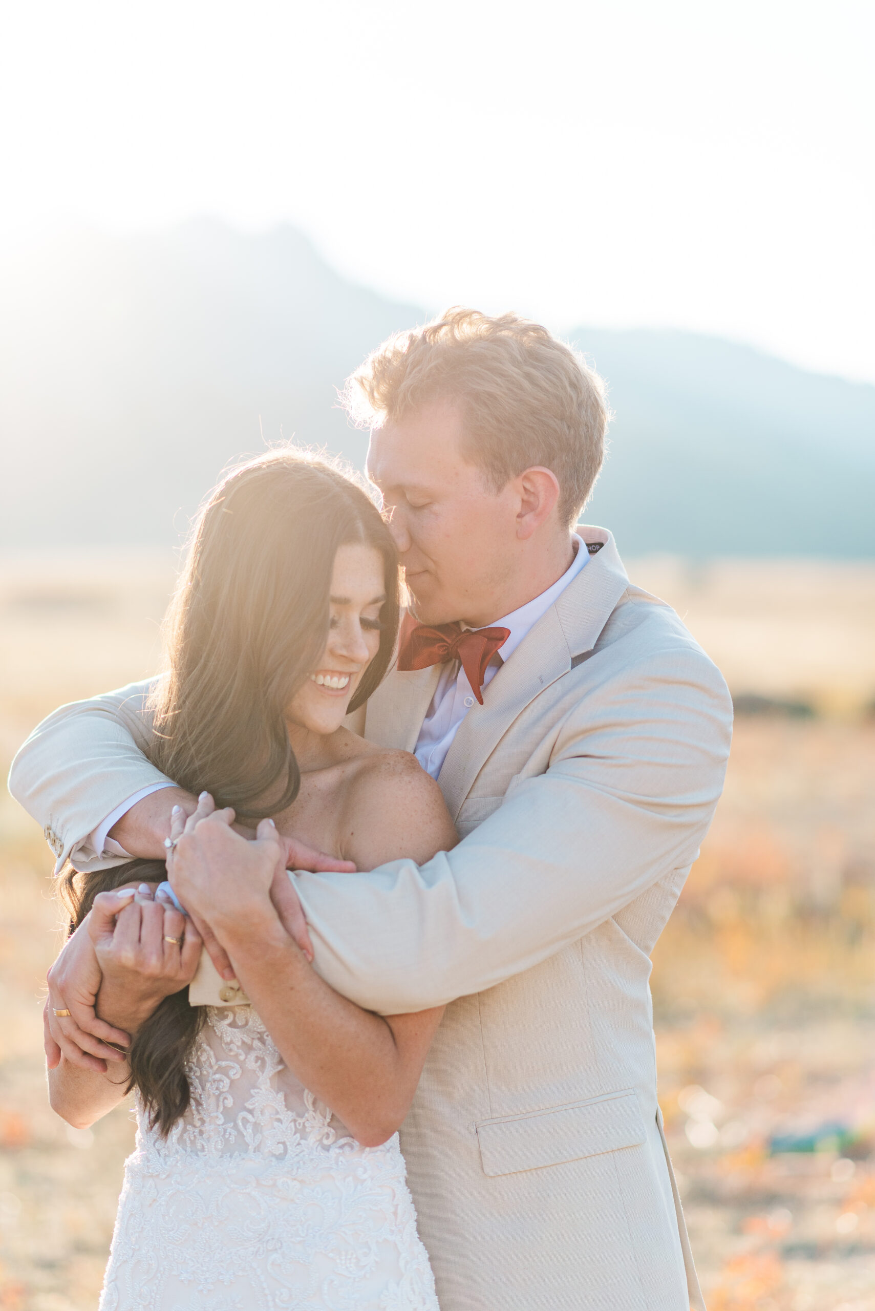 A groom in a tan suit hugs his bride from behind as he places his nose on her temple and they both close their eyes and smile while standing in a field with mountains in the background with Colorado wedding planners help.