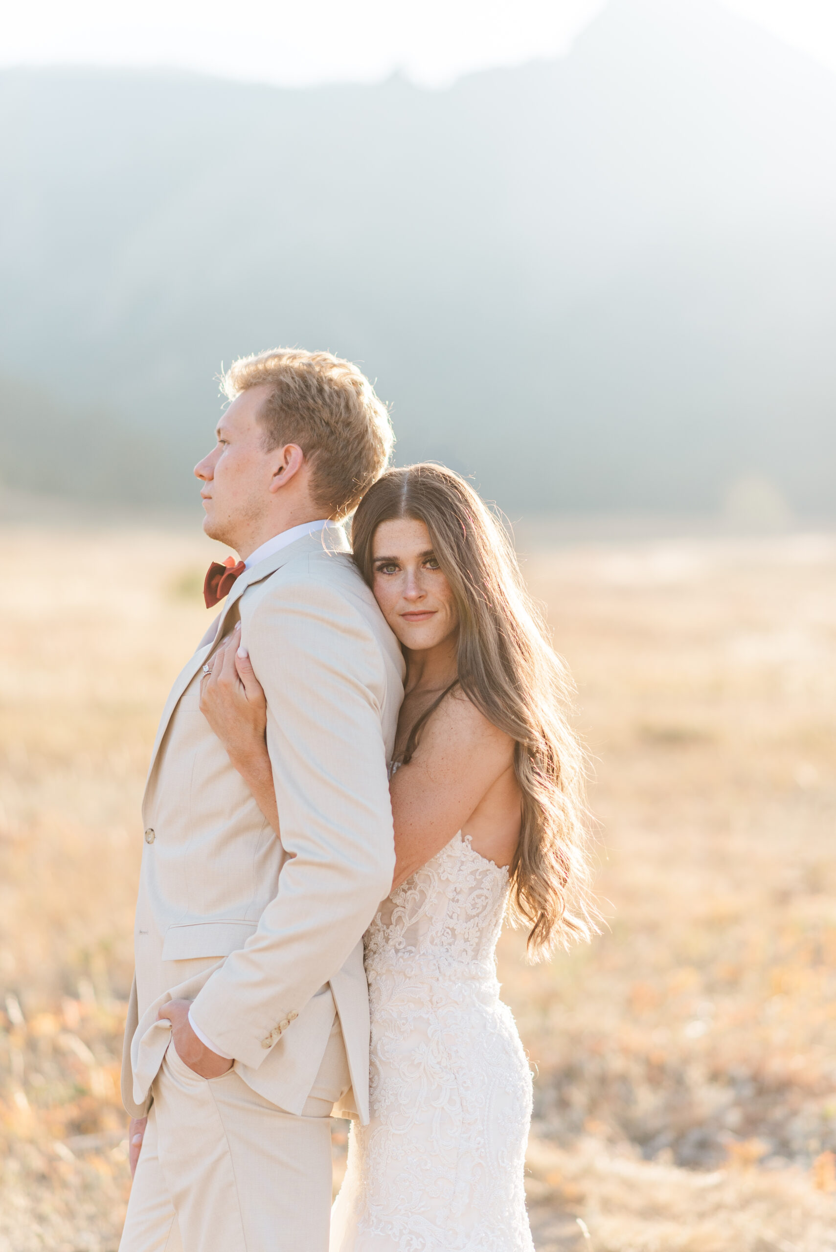 A bride hugs her groom from behind while he stands with a hand in his pocket looking off frame and the bride gives a soft smile toward the camera. 