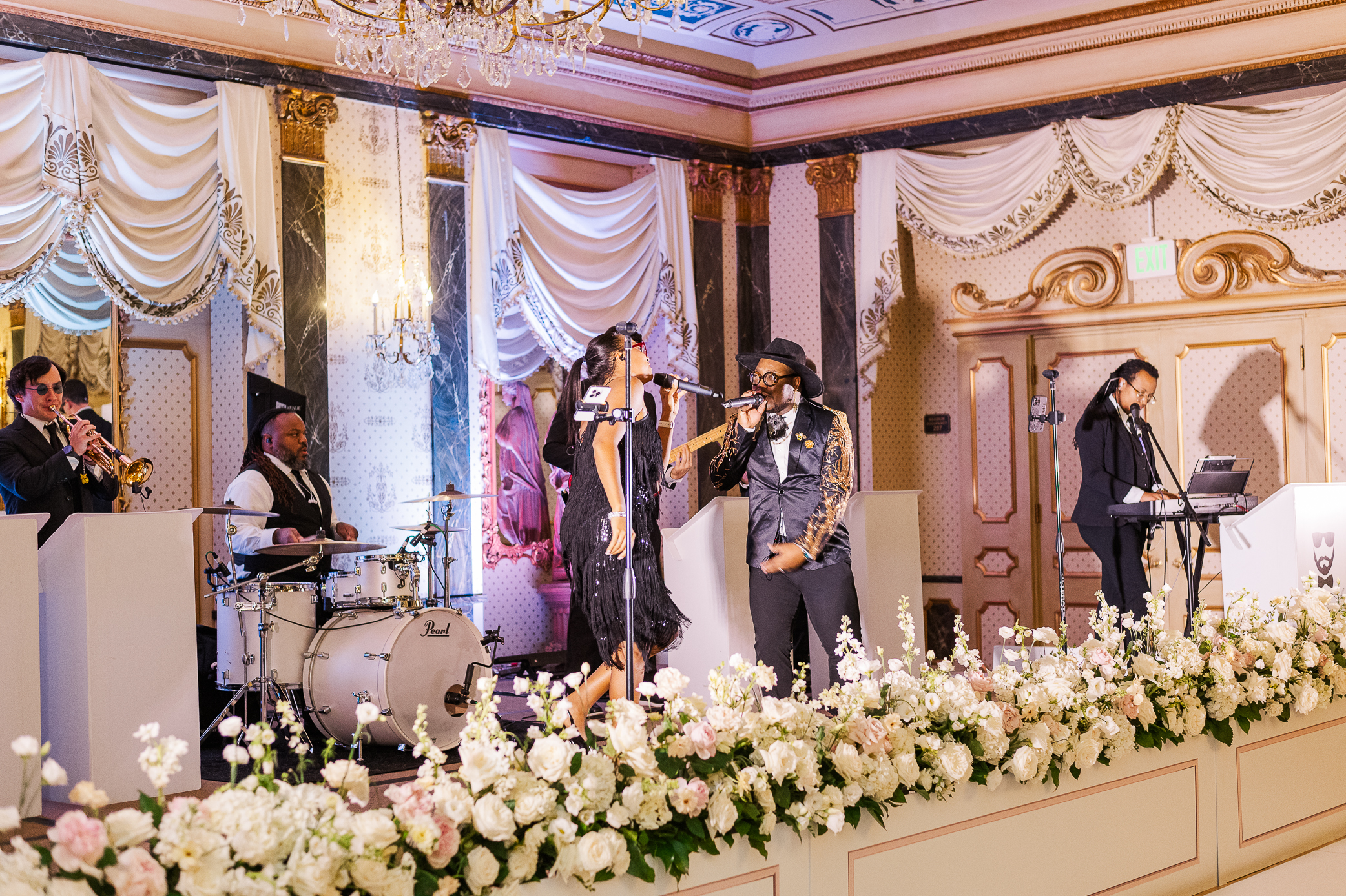 A couple in a pop band sings for a wedding reception. White flowers line the stage in the Spanish colonial revival style room