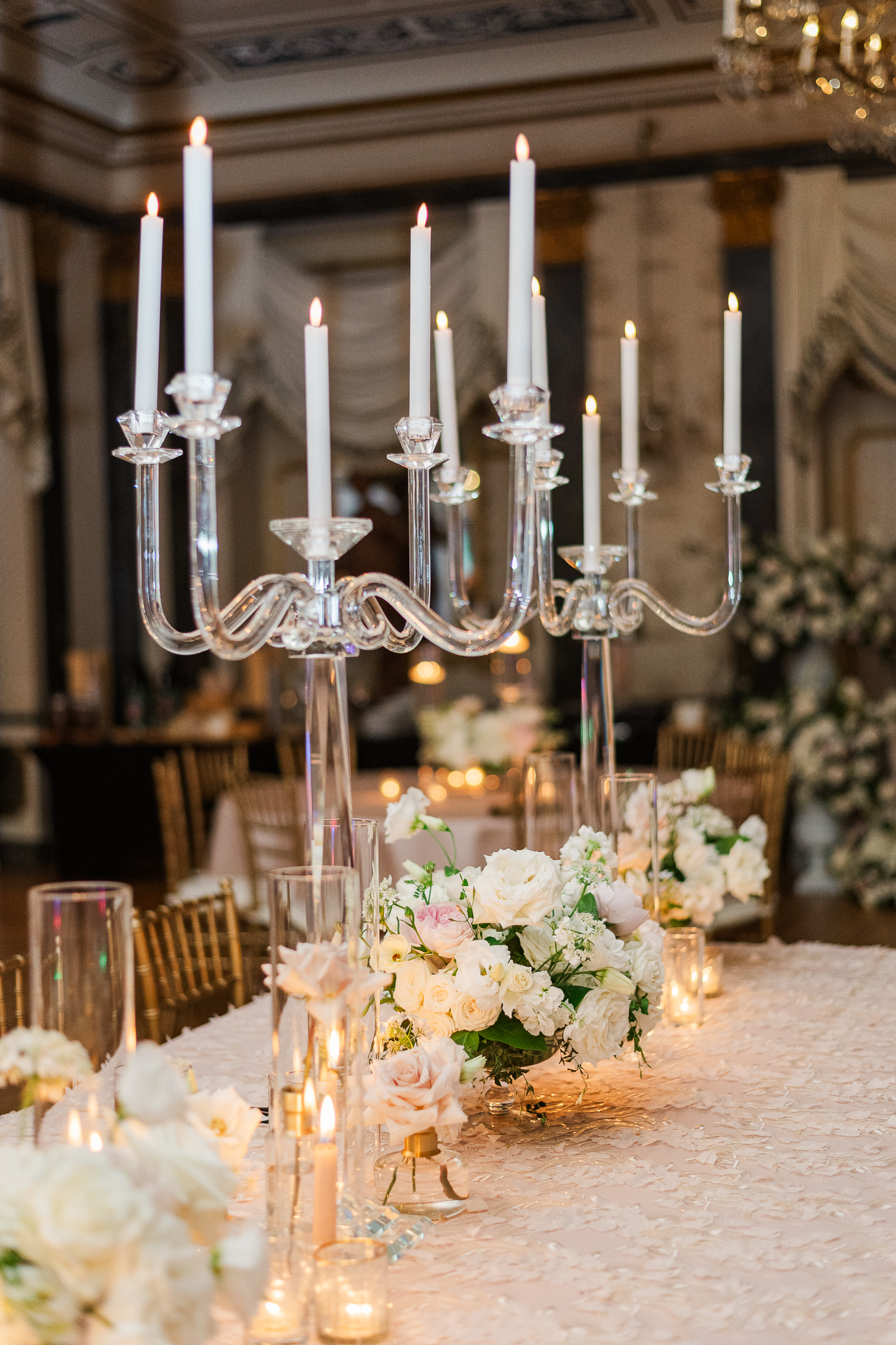 A wedding reception table set up consisting of large clear candelabras, white and pink florals and an extravagant light pink and lace table cloth.