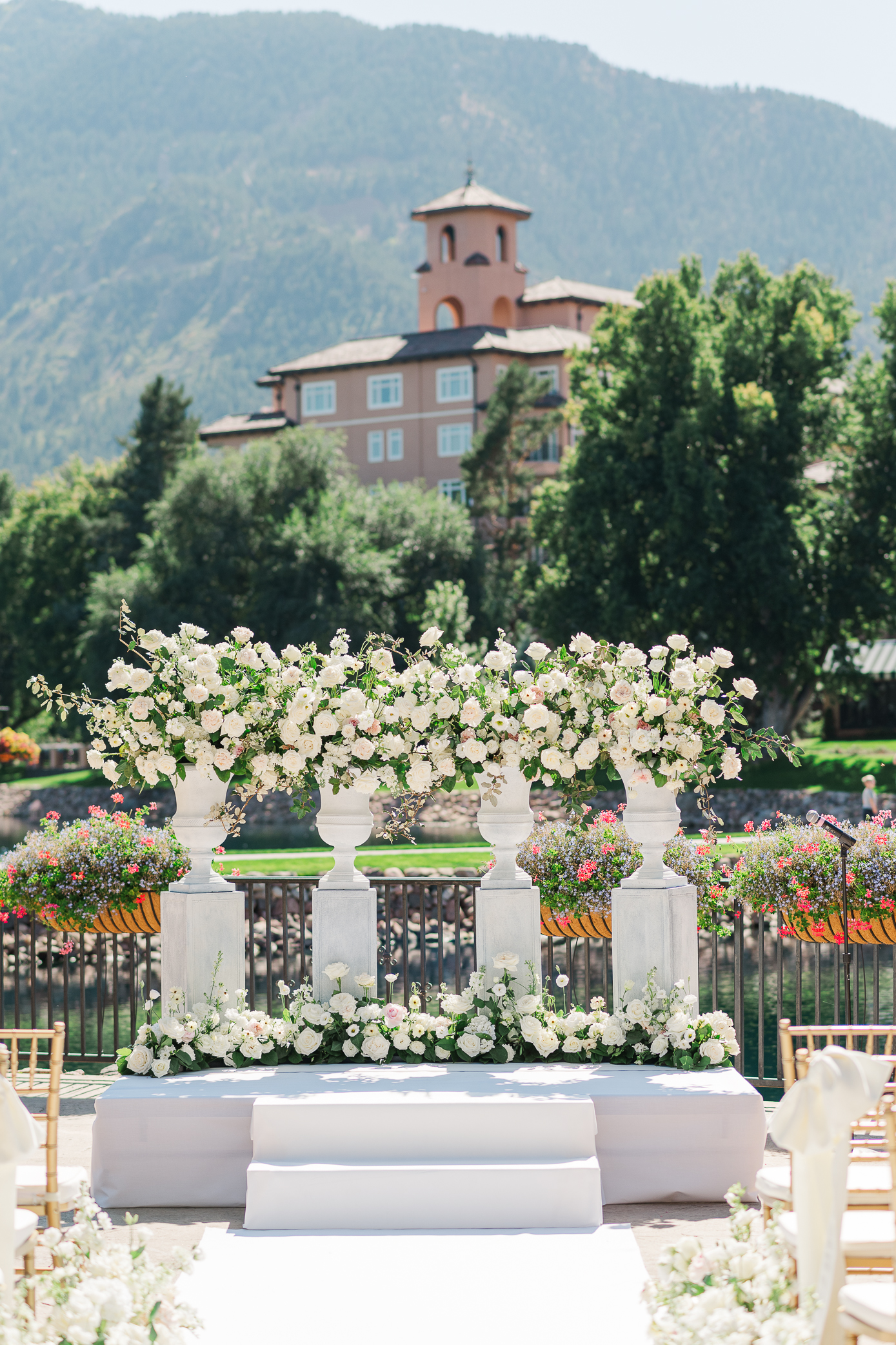 A close up of the alter for a Broadmoor wedding with a white stage, white pillars, and white and pink flowers with a view of Cheyenne mountain and a building a part of the Broadmoor in the background