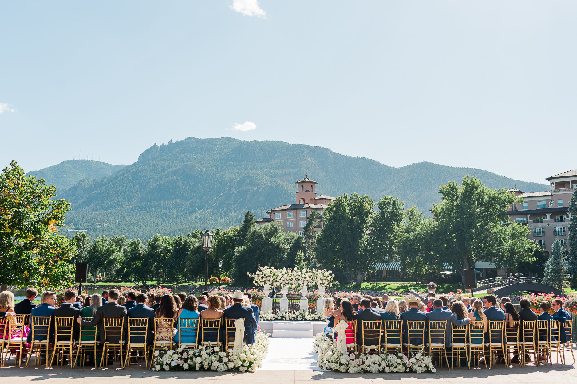 A Broadmoor wedding ceremony set in the heart of Rocky mountains with guests sitting in brown wooden chairs and florals lining the isle and the alter. A beautiful view of Cheyenne mountain with buildings in the background can be seen from the perspective of the guests.