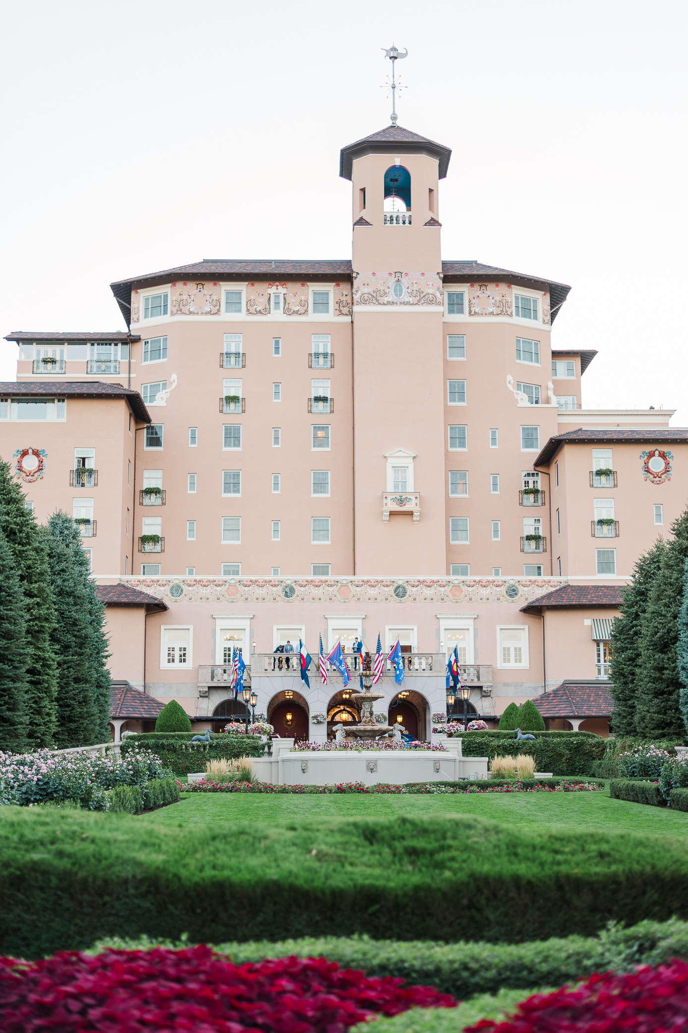 The exterior of the Broadmoor hotel with a well manicured lawn and florals with the exterior of the hotel building in a style of Spanish Colonial Revival 