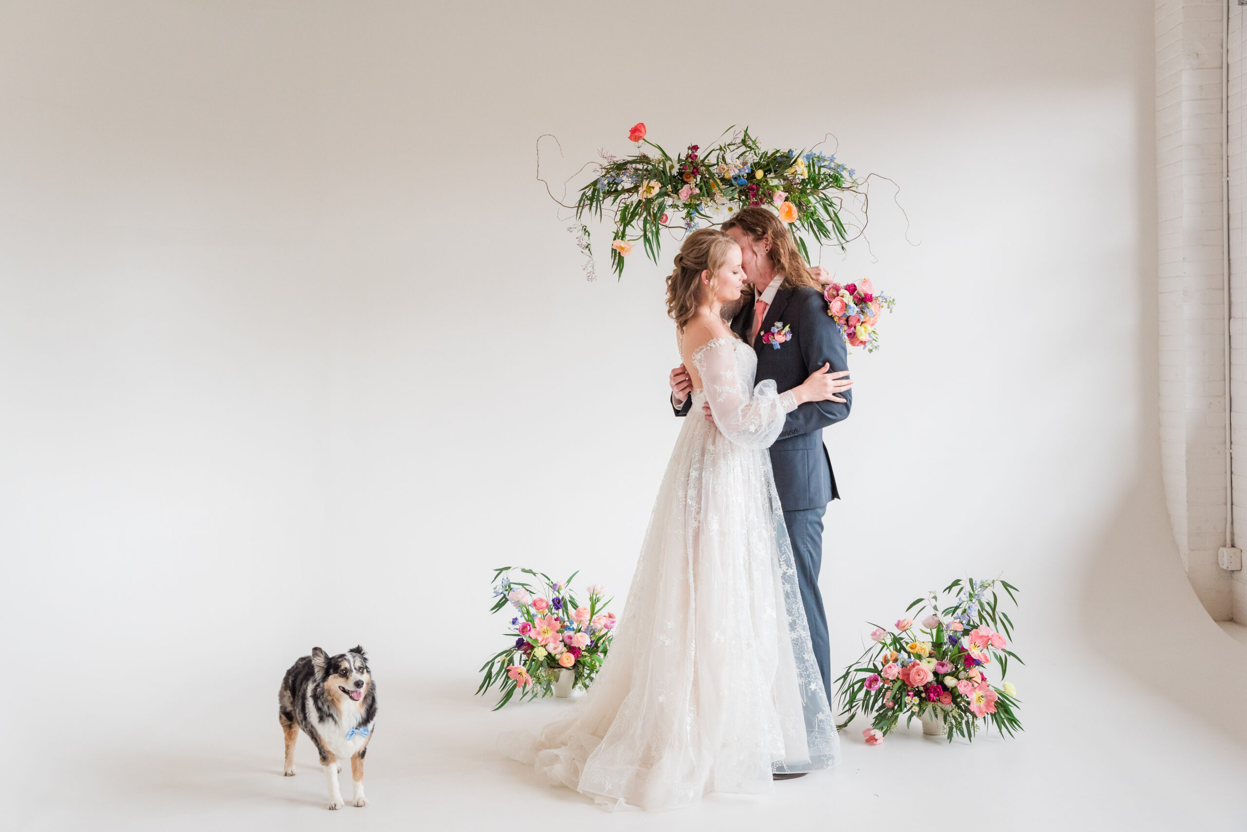 Colorado wedding planners help create a styled shoot at Realm Denver with a whimsical wedding dress on a bride embracing her groom in a grey suit under a whimsical and colorful hanging floral piece with a dog standing next to them