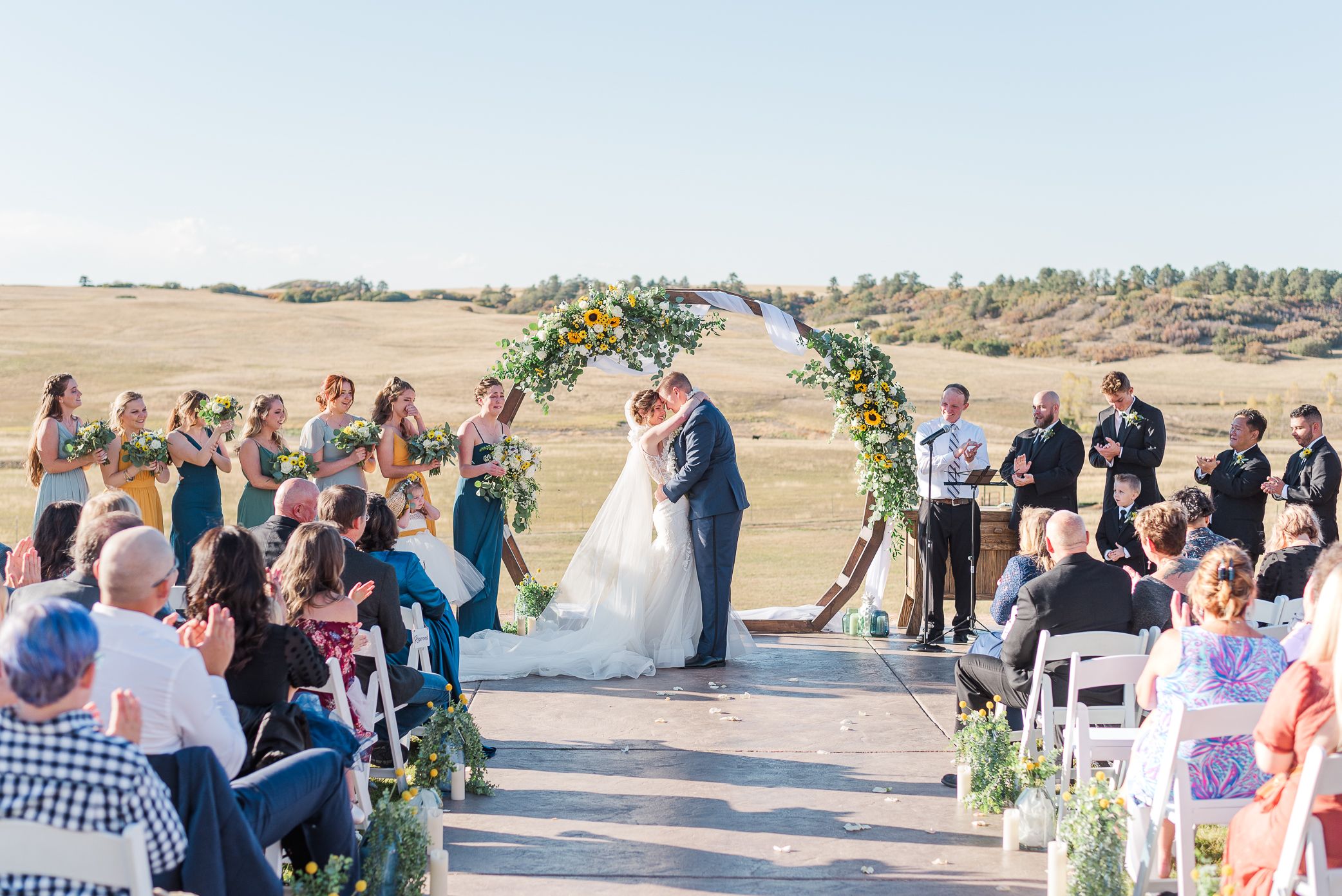 A couple embraces for their first kiss during their wedding ceremony with a round arch in the background and greenery around it. Guests look on and cheer.