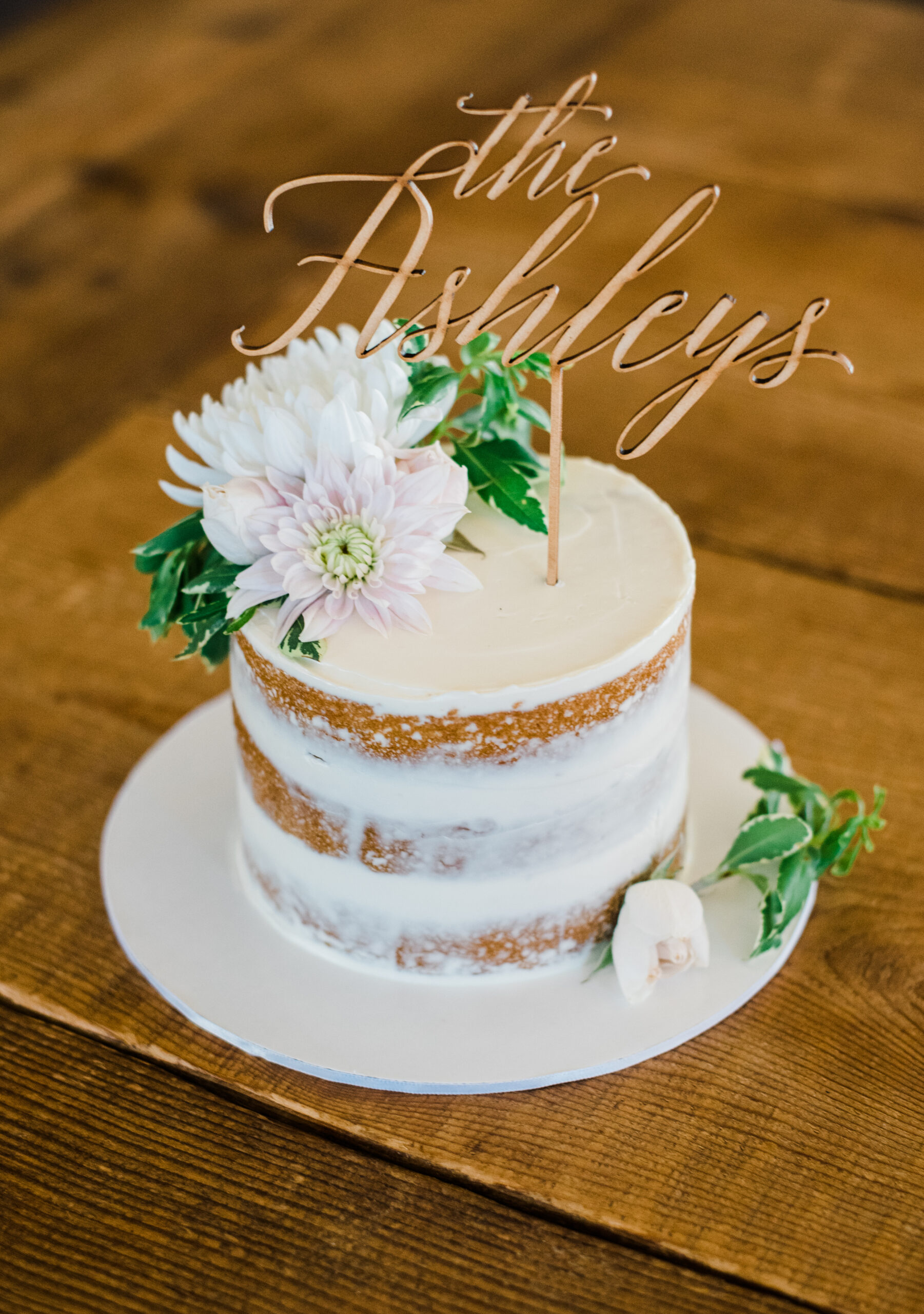 A close up of a wedding cake with white frosting and several flowers on the top of the cake with a sign that says "the Ashleys"