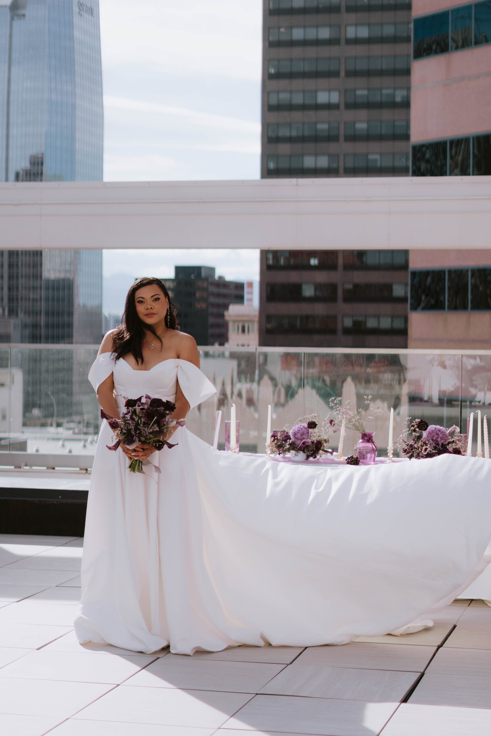 An image of Weddings by Jay in a bridal gown in front of the dessert table. The woman is hold a bouquet in front of her with purple flowers