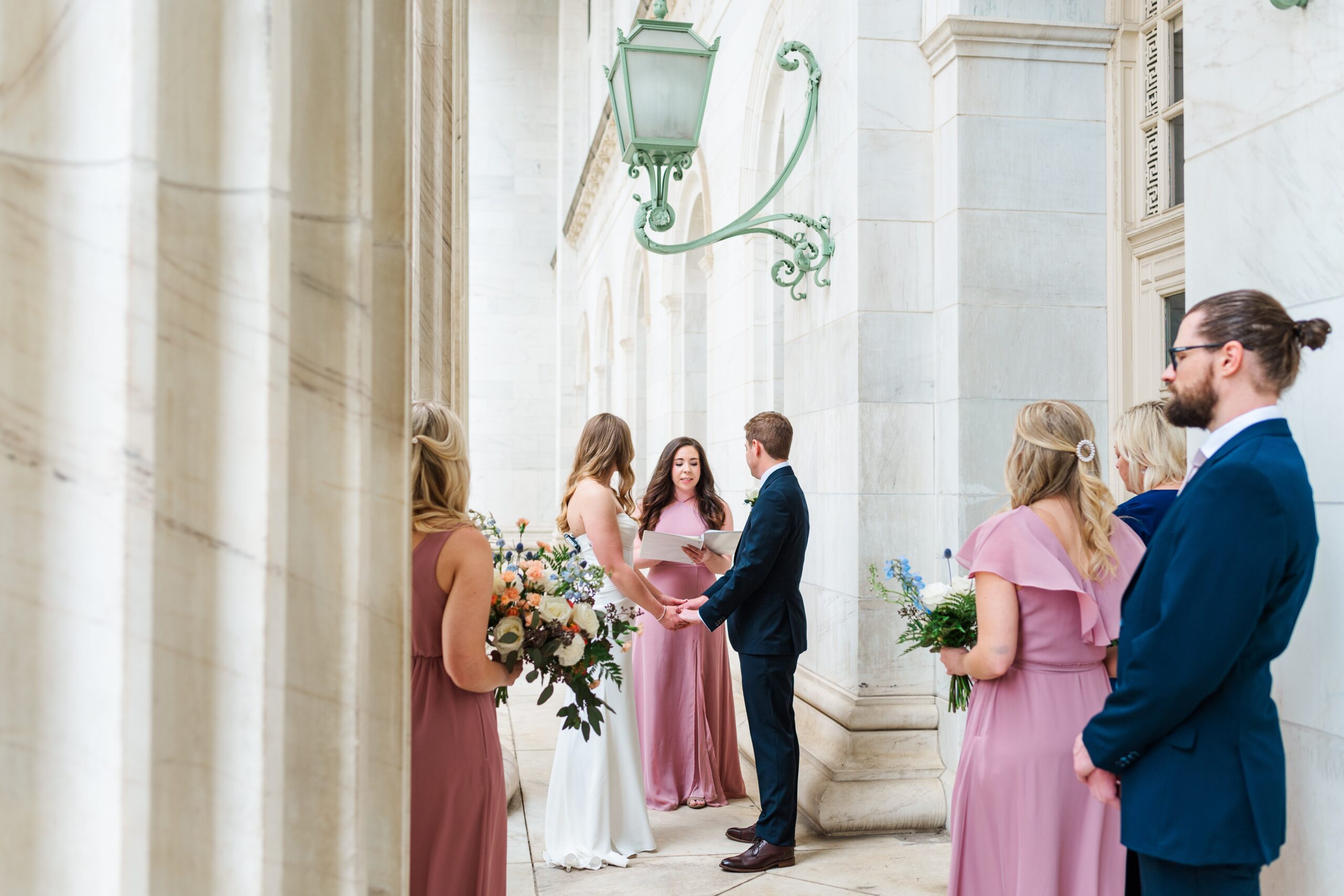  Byron White Courthouse wedding with a woman in a pink dress officiating for the bride in a white dress and the groom with a navy blue suit. Family members stand close by in pink dresses and navy suits while the bridesmaids hold flower bouquets.