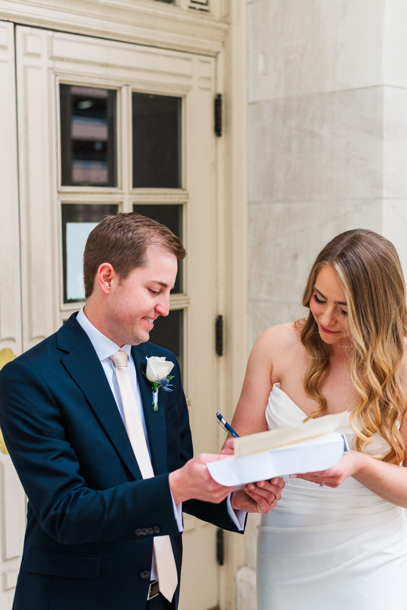 A bride stands next to her new husband and smiles as she signs the marriage license that the groom is holding for her immediately following their Byron White Courthouse wedding