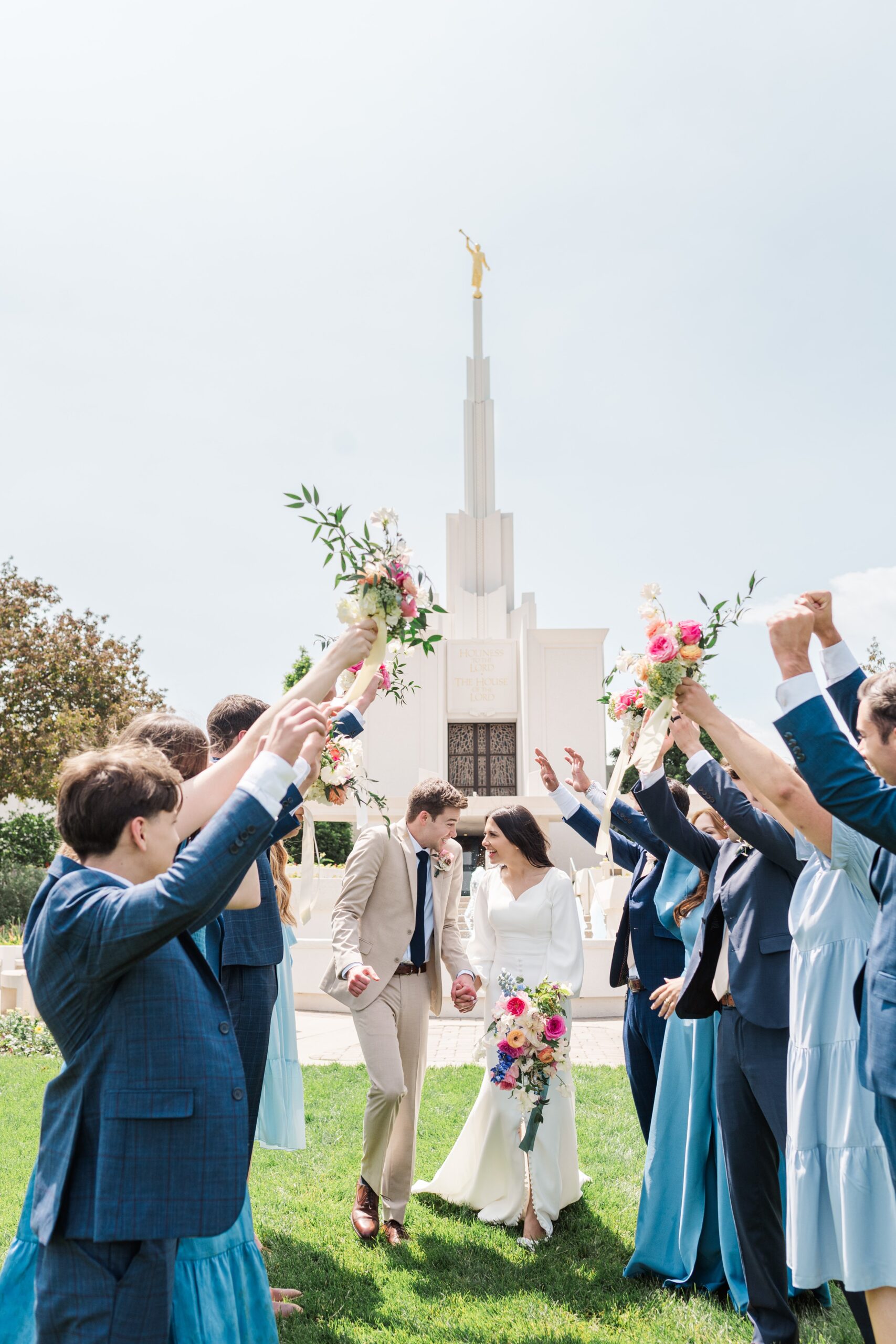 Bride and groom at the Denver LDS Temple wedding walk through a tunnel made by their friends standing in a line and hold their hands up as they walk through. 